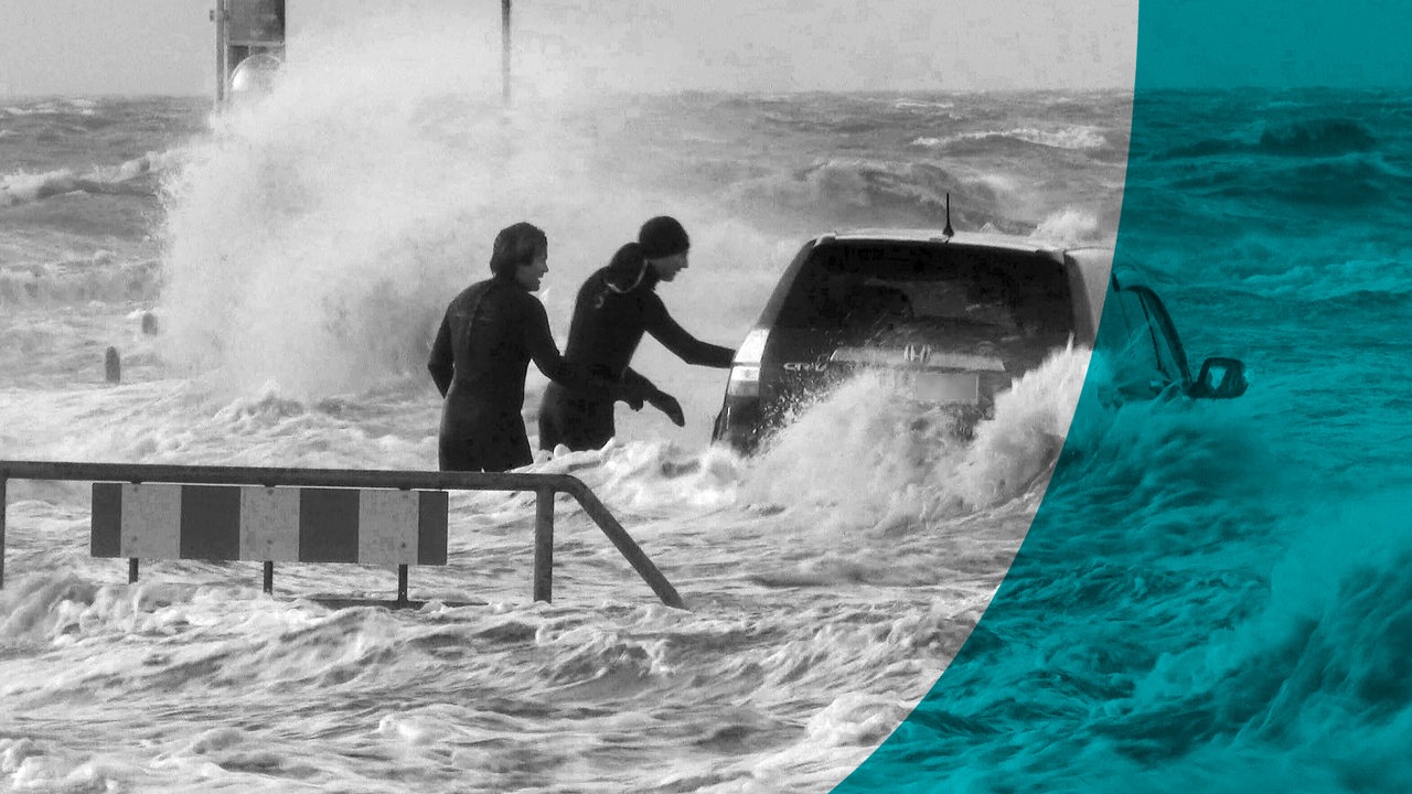 Ein Auto steht auf einem Parkplatz der Mole des Fähranlegers im Wasser der Nordsee. Zwei Kitesurfer schauen in das Fahrzeug eines Unbekannten.