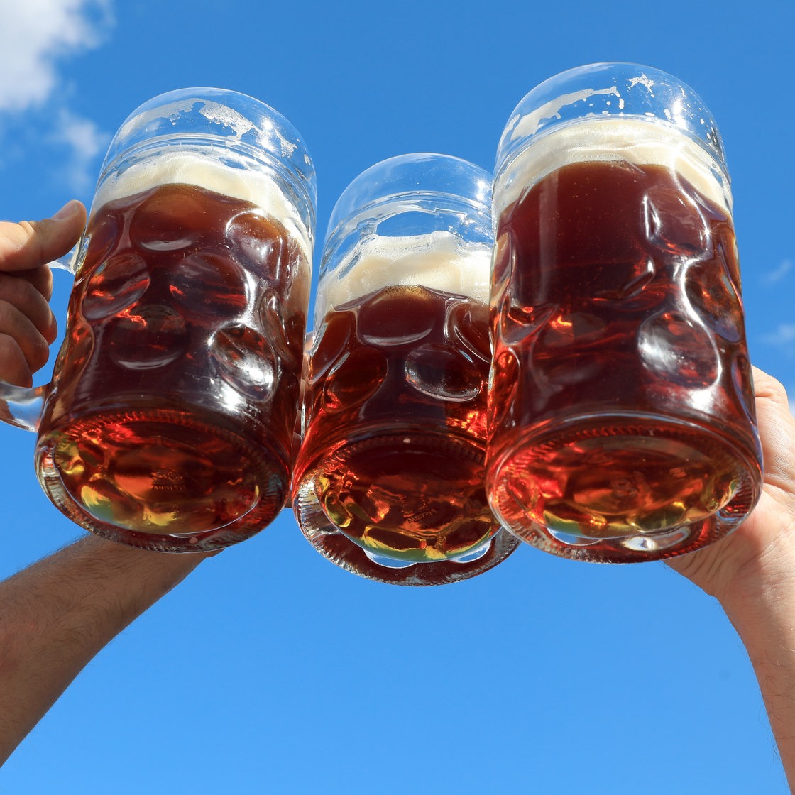 Drei Hände stoßen gegen den blauen Himmel mit gläsernen Bierkrügen an.