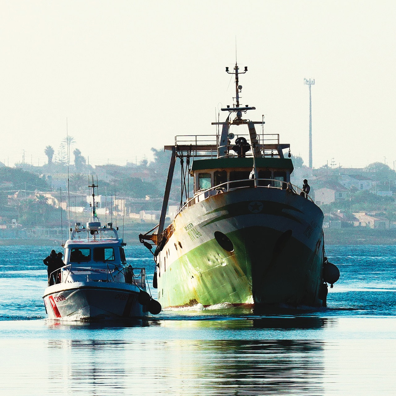 Ein Schiff wird begleitet von einem Polizeischiff. Im Hintergrund sieht man eine Hafenstadt.