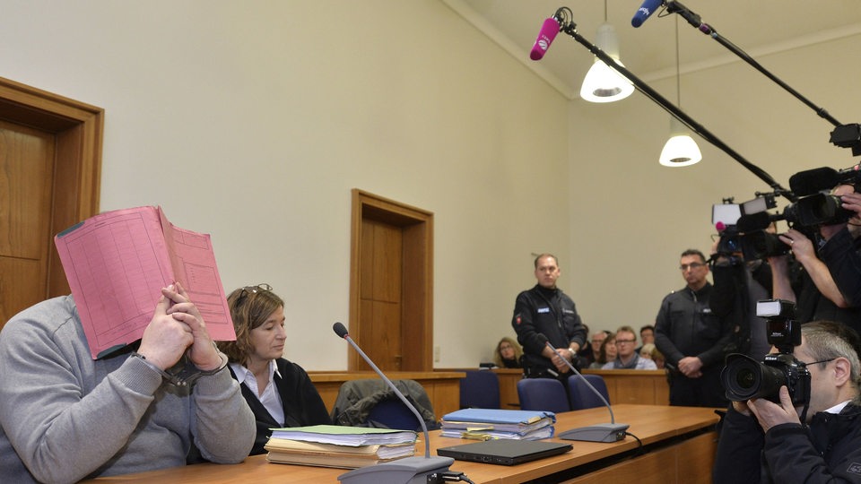 Niels H. sitzt vor Gericht auf der Anklagebank und hält sich eine Mappe vor das Gesicht und ist von Pressevertretern umringt.