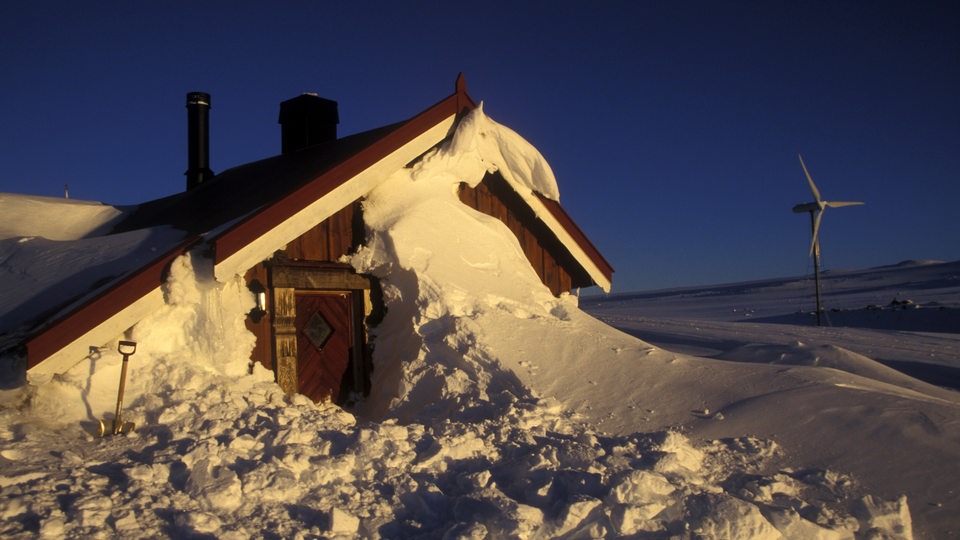 Ein eingeschneites Haus, daneben eine Windkraftanlage (Symbolbild)