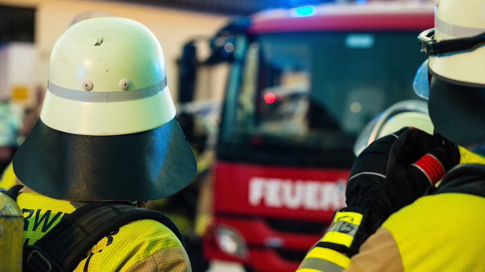 Blick von hinten auf zwei Feuerwehrmänner mit Schutzausrüstung, dahinter unscharf die Front eines Feuerwehrfahrzeugs mit leuchtendem Blaulicht (Symbolbild)