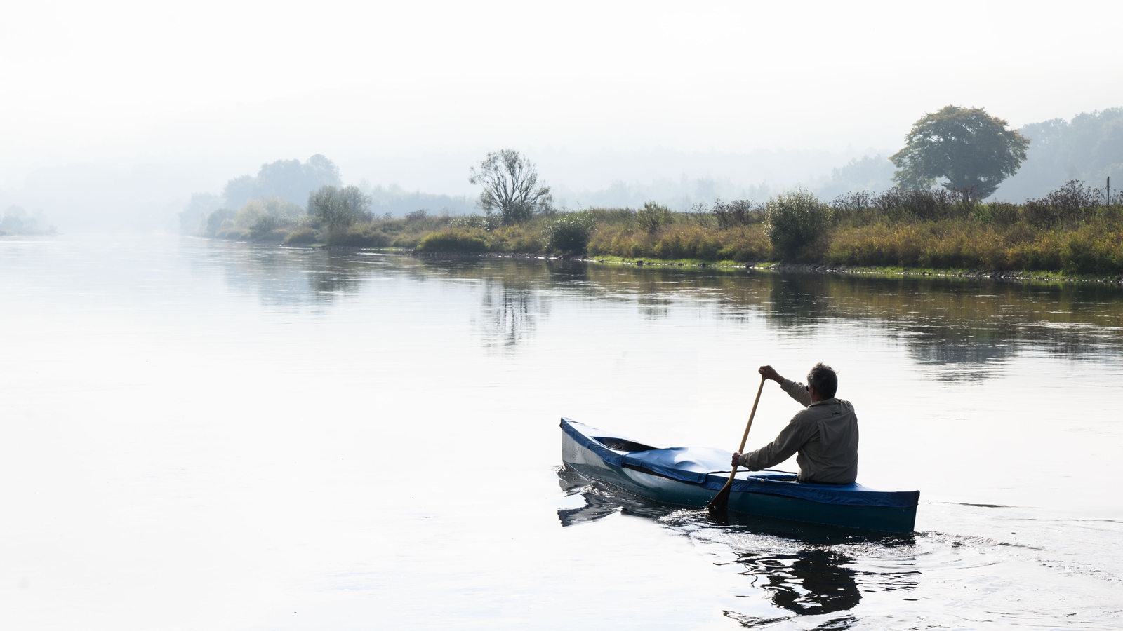 Ein Mann paddelt im Morgennebel über einen Fluss