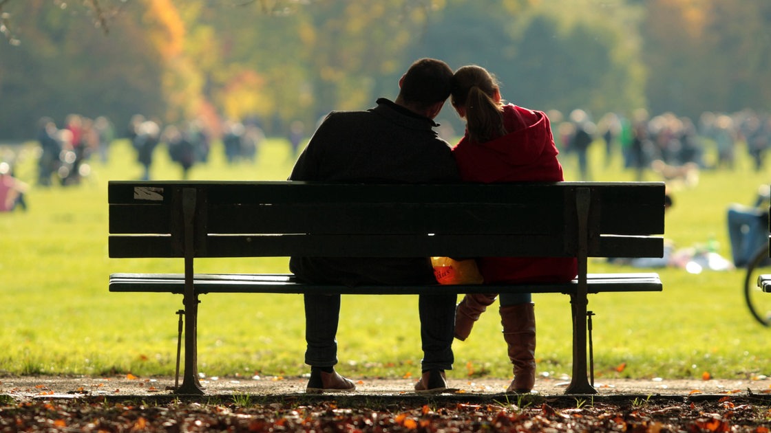 Ein junges Paar in Rückenansicht auf einer Parkbank schaut auf eine Wiese mit vielen Menschen