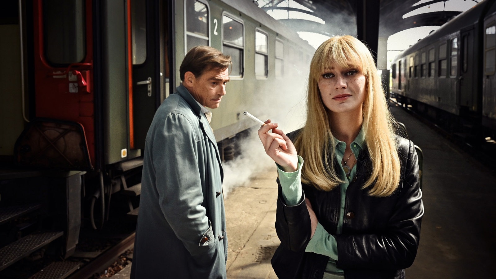 Ein Mann blickt misstrauisch auf eine Frau mit Zigarette auf einem Bahnsteig zwischen zwei Zügen.