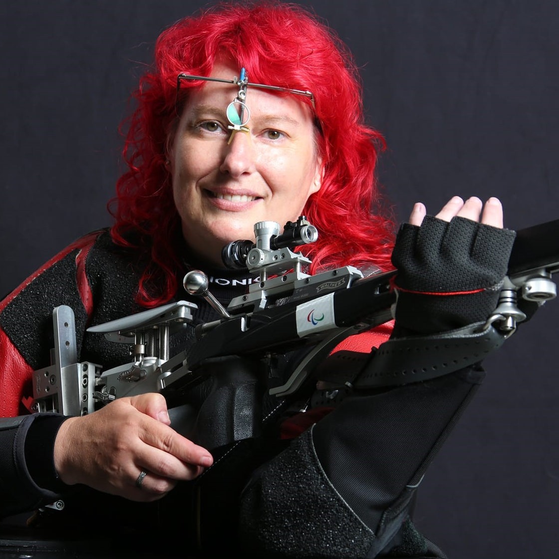 Elke Seeliger in schwarz-roter Schießmontur und mit Luftgewehr, mit rot gefärbten Haaren vor schwarzem Hintergrund