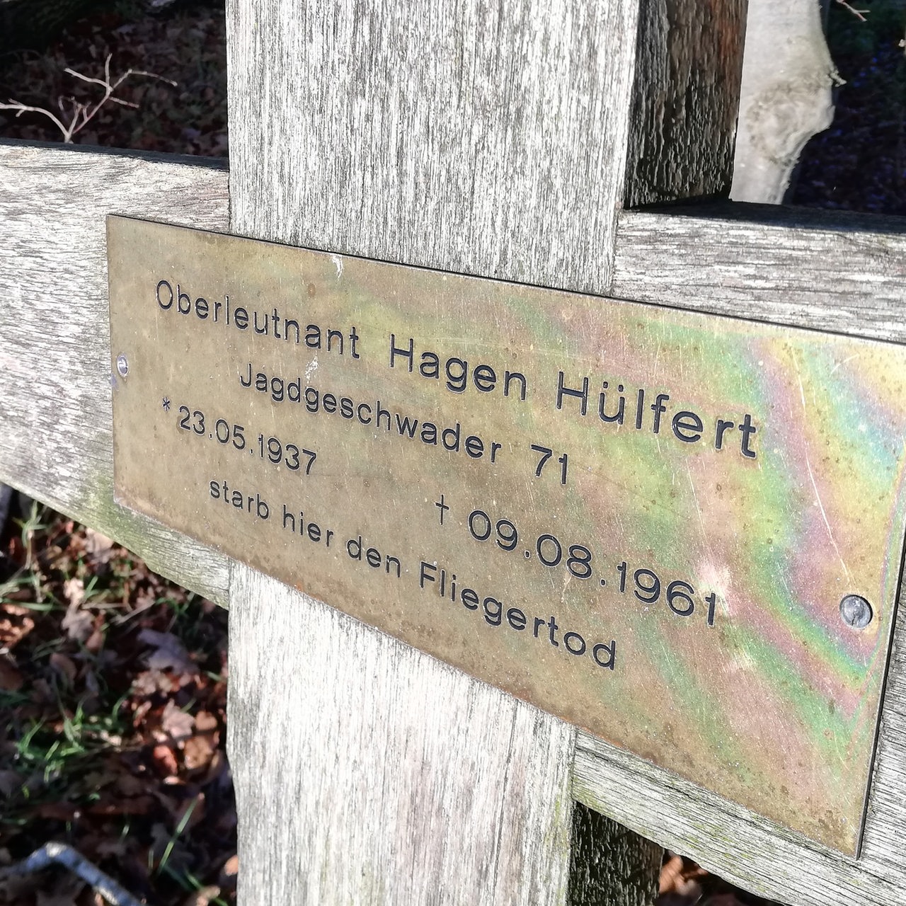 Eine Namensplakette auf einem Holzkreuz zwischen Bäumen.