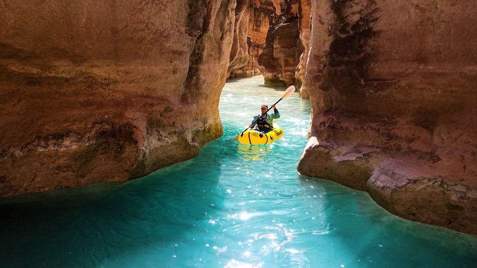 Eine Frau paddelt zwischen rot-gelben Felsformationen des Grand Canyons in blauem Wasser