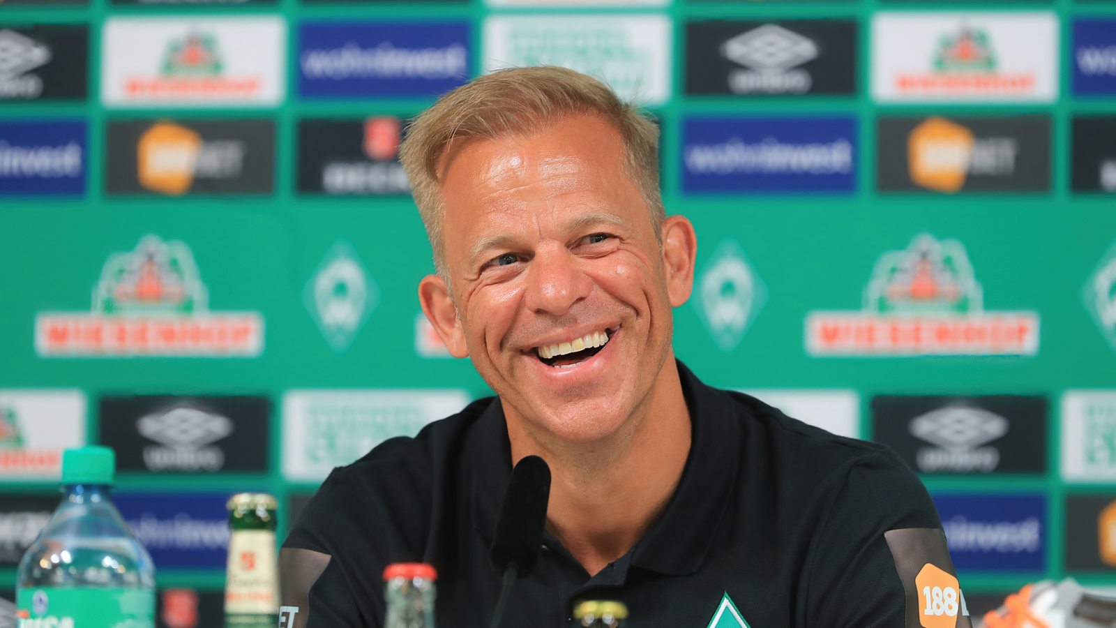 Trainer Markus Anfang strahlend bei seiner offiziellen Vorstellung bei Werder Bremen vor einer Werbewand.