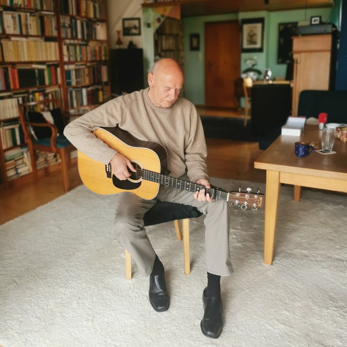 Ein Mann in einem beigen Pullover spielt Gitarre in einem Wohnzimmer