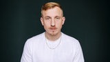 Hubertus Koch steht im weißen T-Shirt vor einer dunklen Wand und schaut in die Kamera