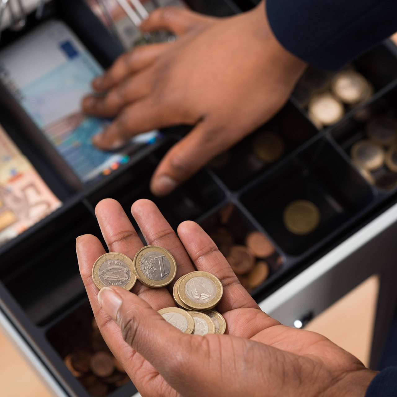 Zwei Hände greifen in die Kassenschublade einer Supermarktkasse.