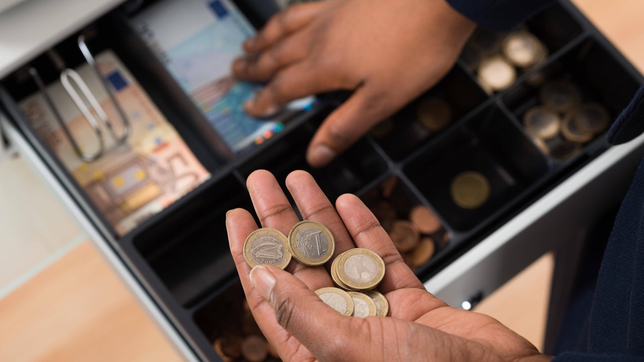 Zwei Hände greifen in die Kassenschublade einer Supermarktkasse.