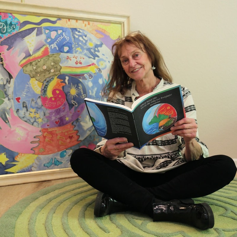 Karin Grabenhorst sitzt im Schneidersitz vor einer Bildcollage und hält ein Buch in der Hand.