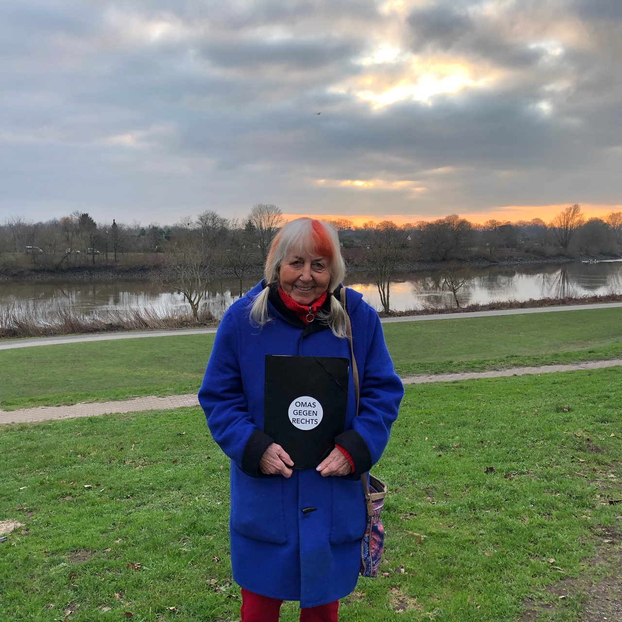 Gerda Smorra steht auf einer Wiese und hölt eine Mappe mit der Aufschrift "Omas gegen Rechts" in der Hand