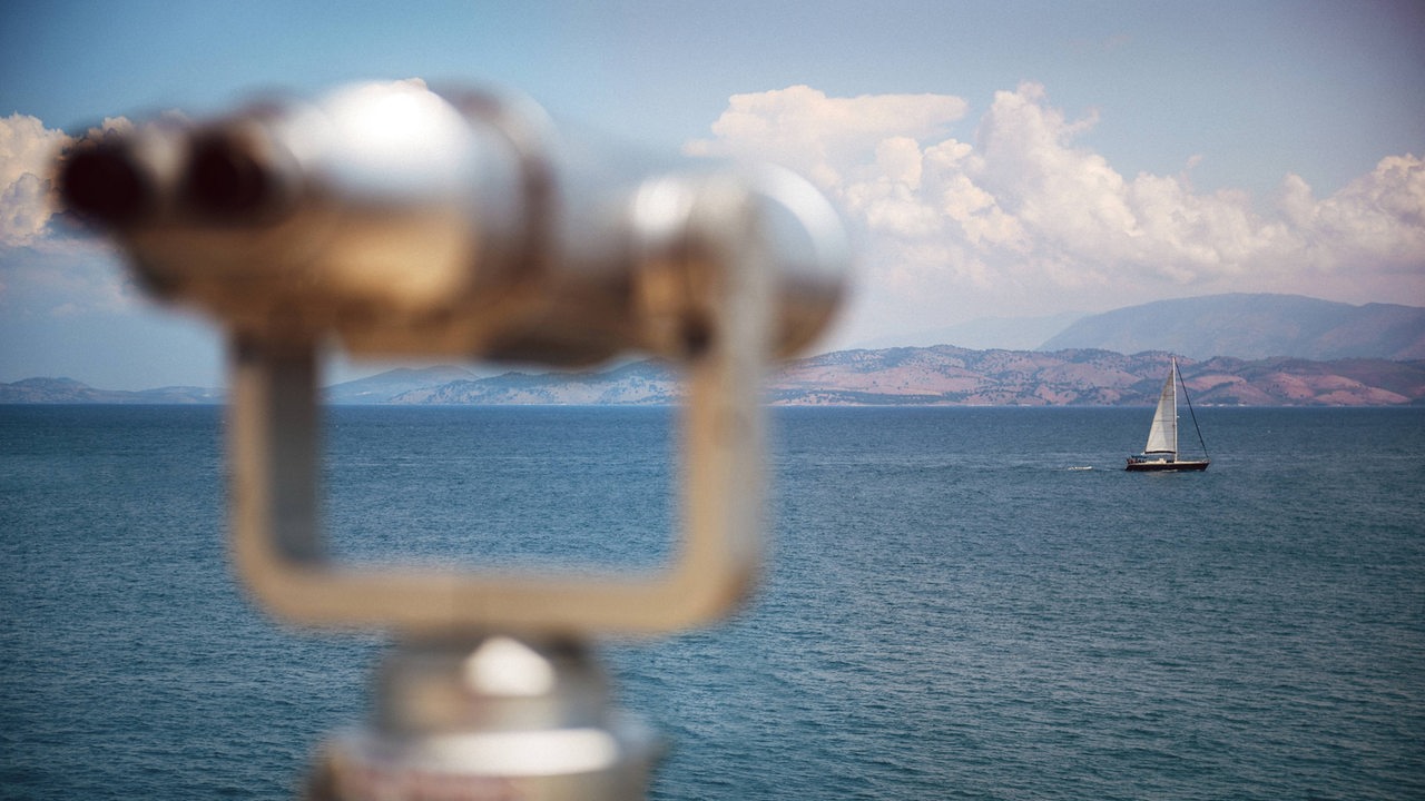 Ein Meer mit Segelboot, im Hintergurnd Küste, im Vordergrund ein unscharfes Fernglas (Symbolbild)