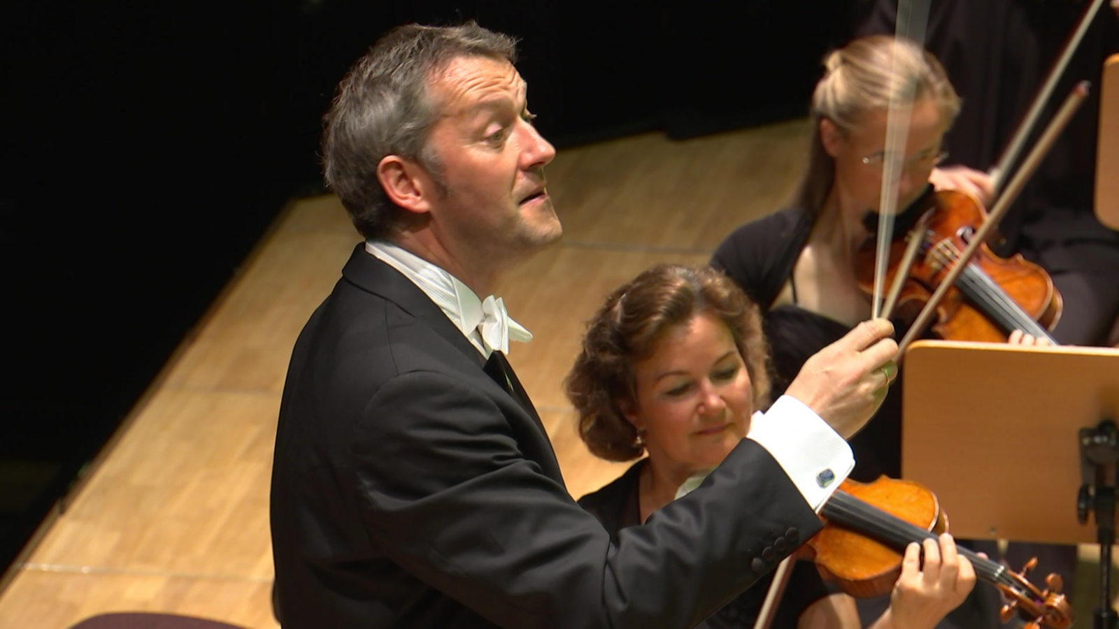 Dirigent Markus Poschner dirigiert bei einem Konzert, im Hintergrund zwei Geigerinnen.
