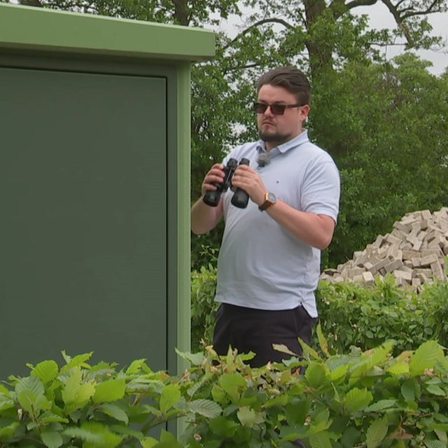 Privatdetektiv Michael Günther versteckt mit Sonnenbrille und Fernglas in der Hand, hinter eine Hecke. 