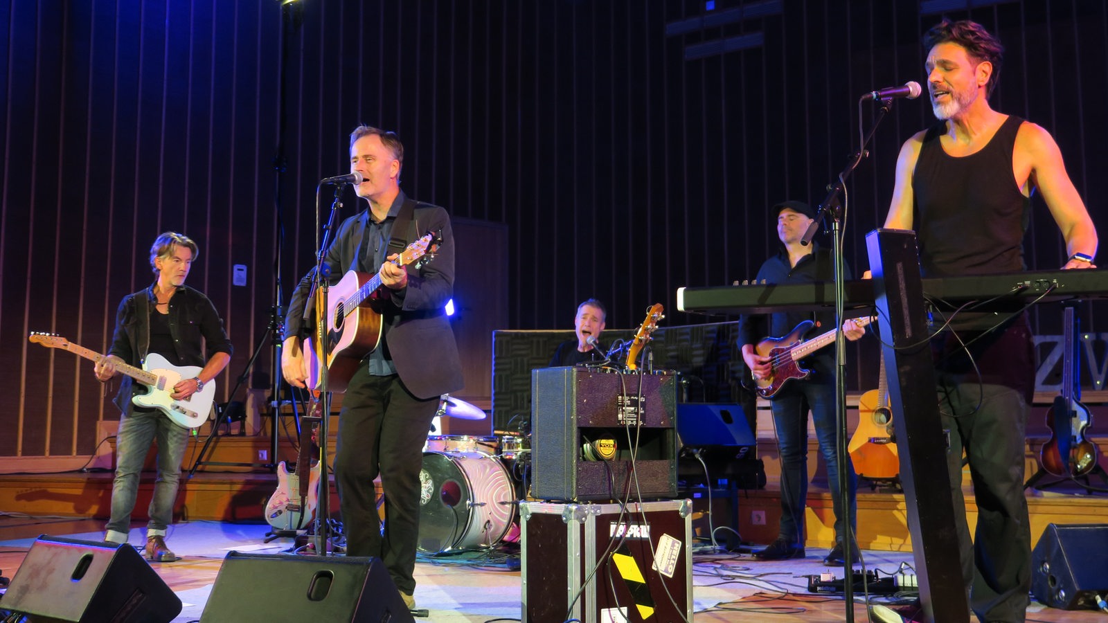 Fünf Männer mit Instrumenten stehen auf einer Bühne.