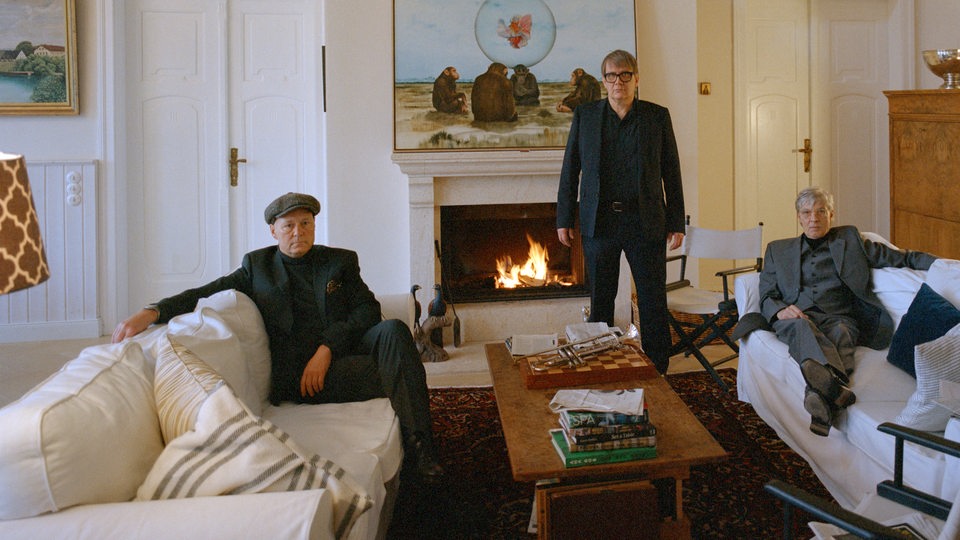 Drei Männer stehen und sitzen in einem hellen Zimmer