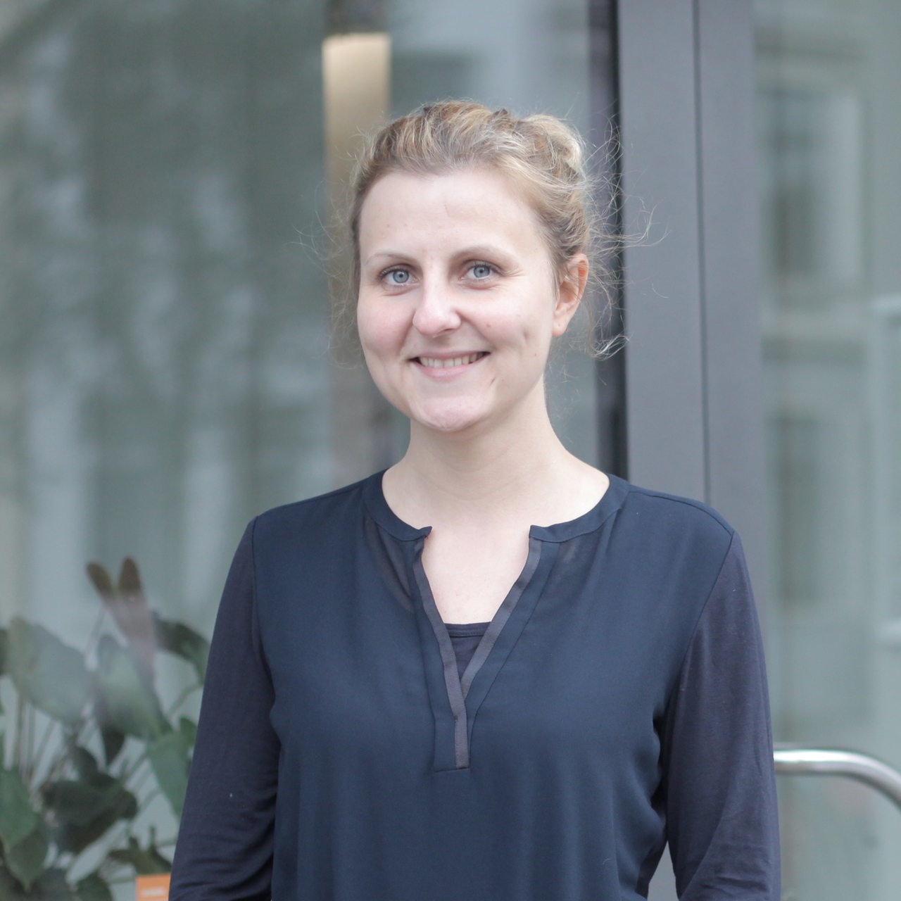 Eine blonde junge Frau in einem blauen Shirt steht vor einem Fenster