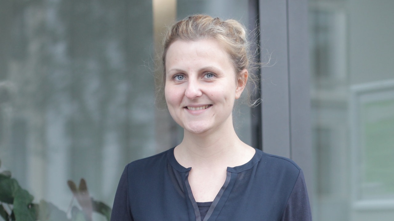 Eine blonde junge Frau in einem blauen Shirt steht vor einem Fenster