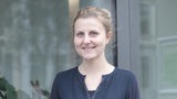 Eine blonde junge Frau in einem blauen Shirt steht vor einem Fenster