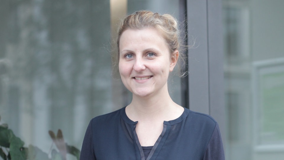 Eine blonde junge Frau in einem blauen Shirt steht vor einem Fenster