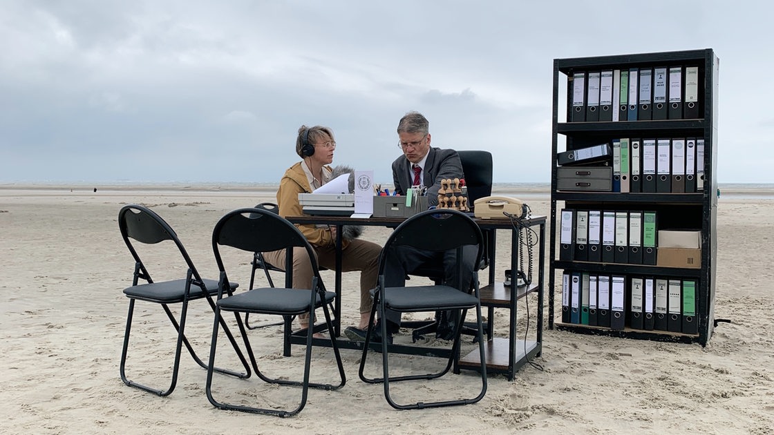 Büromöbel stehen am Strand, eine Frau und ein Mann sitzen am Bürotisch