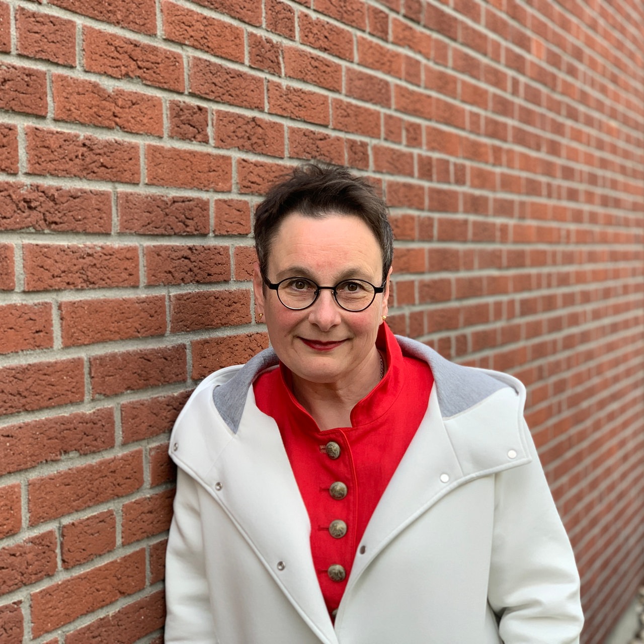 Ein Frau in weißer Jacke steht vor einer Backsteinwand.