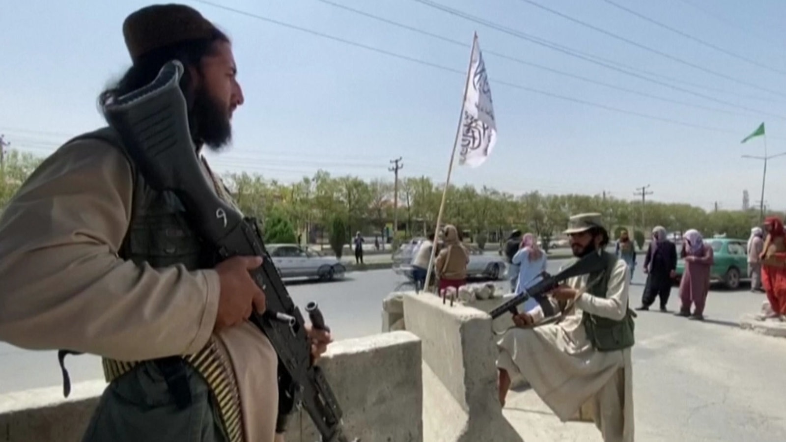 Zwei bewaffnete Taliban stehen an einer Straße in Kabul, nachdem sie die Macht in Afghanistan ergriffen haben. Im Hintergrund sind in unbewaffnete Menschen in traditioneller Kleidung.