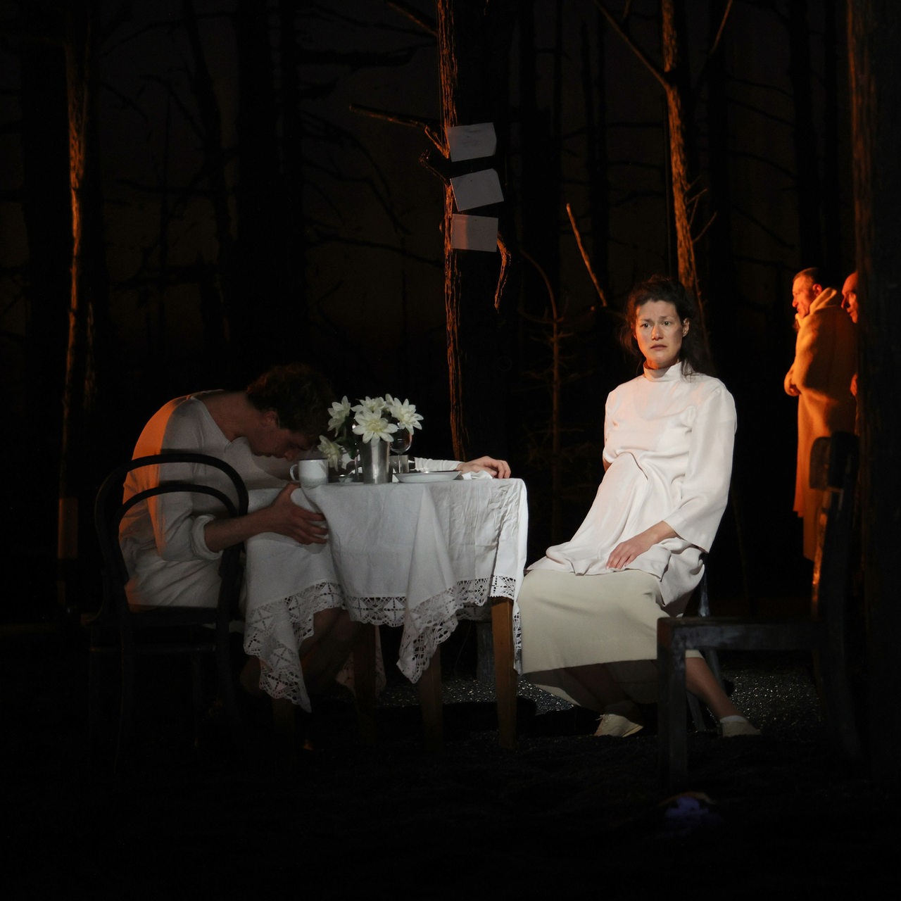 Szene aus "Erbarmen":  Ein Paar sitzt an einem gedeckten Tisch, im Hintergrund sieht man Menschen in einem Wald