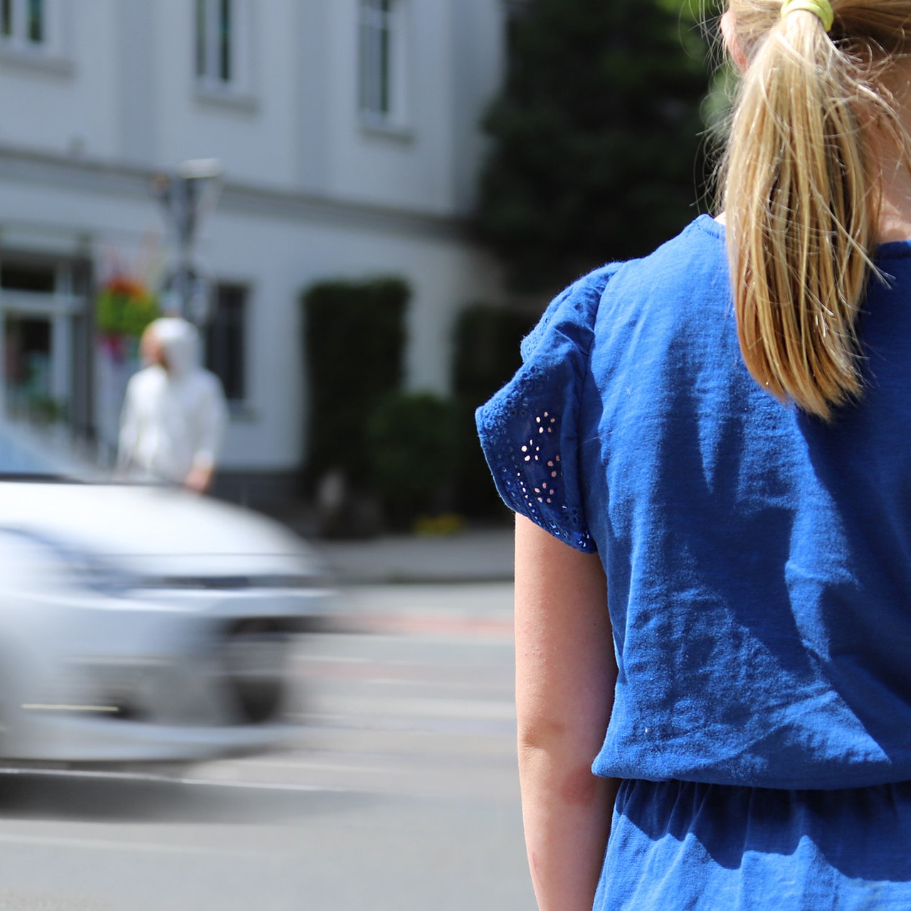 Ein Mädchen im blauen Kleid steht an der vielbefahrenen Kreuzung Friedrich-Ebert-Straße und Neustadtscontrescarpe und versucht über die Straße zu kommen. Im Hintergrund versuchen zwei Erwachsene die Straße zu überqueren.