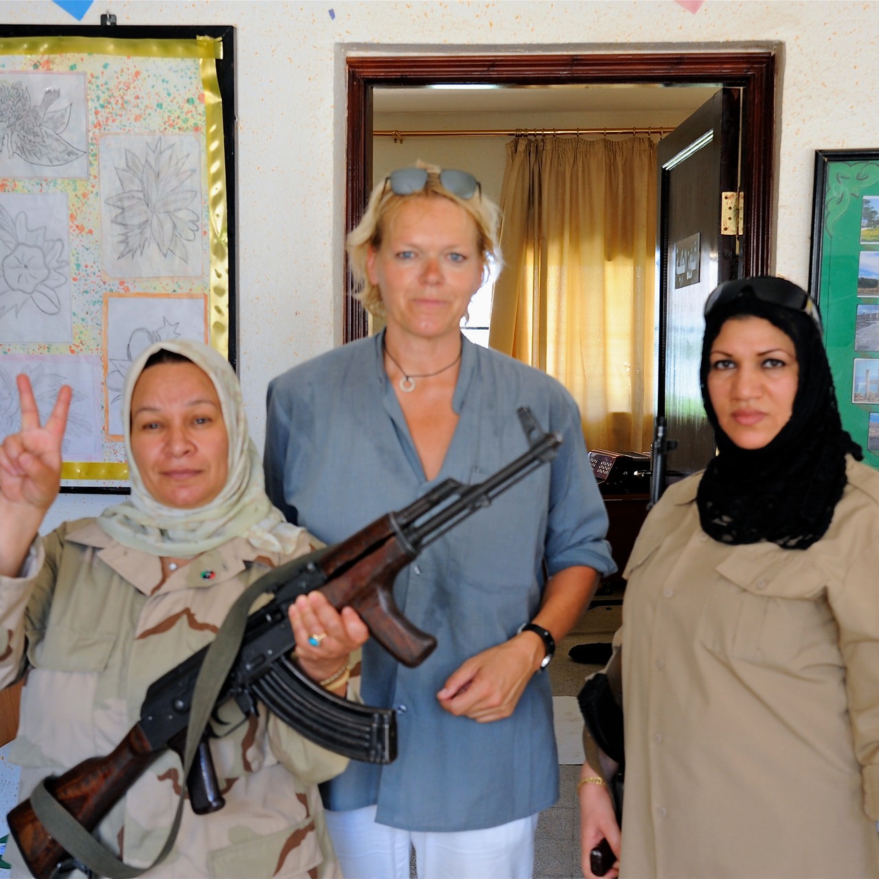 Eine große blonde Frau zwischen zwei dunkelhaarigen Frauen mit Kopftuch. Eine der Frauen trägt ein Gewehr.