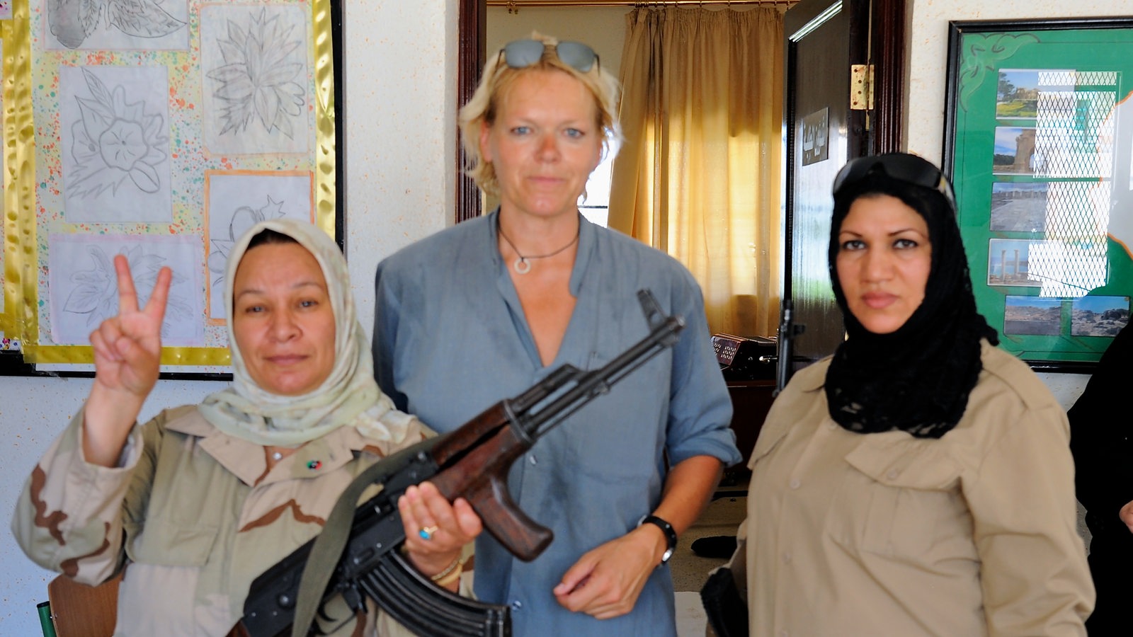 Eine große blonde Frau zwischen zwei dunkelhaarigen Frauen mit Kopftuch. Eine der Frauen trägt ein Gewehr.