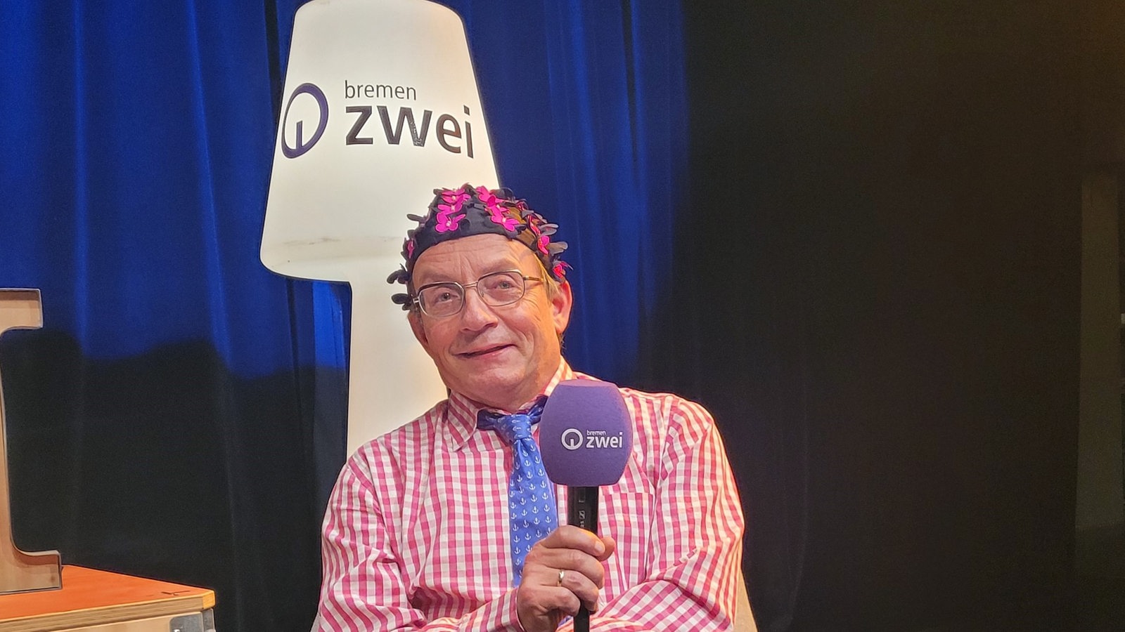 Wigald Boning trägt eine geblümte Badekappe und sitzt mit Mikrofon in der Hand auf der Bremen Zwei Herbstgast-Bühne. 
