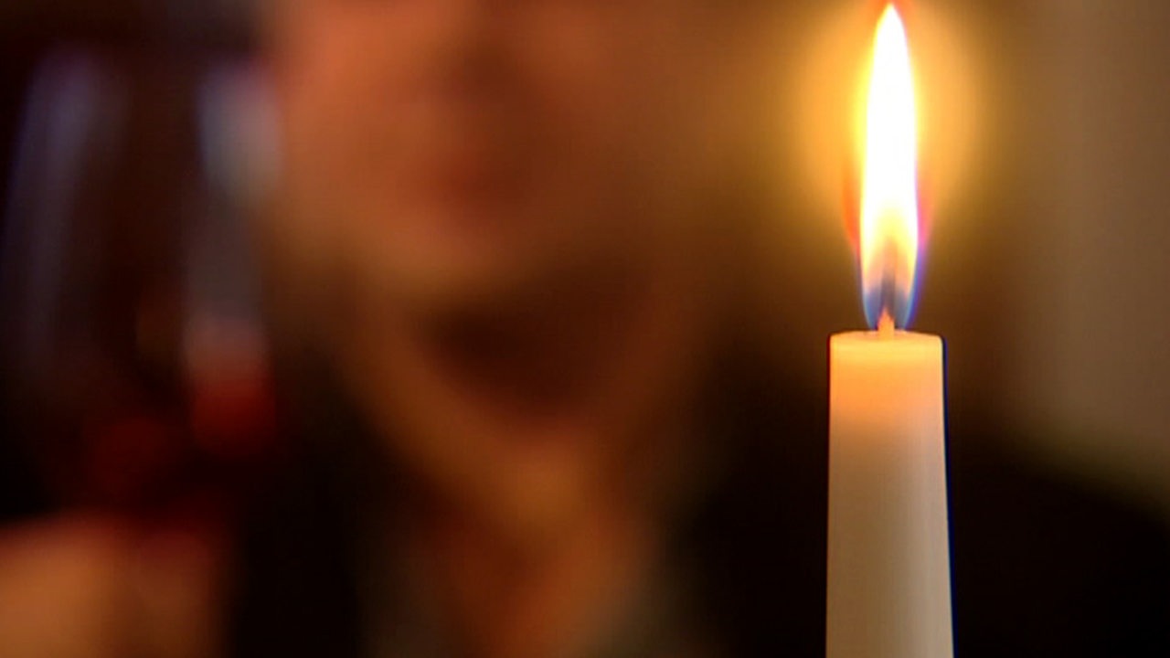 Eine Kerze, im Hintergrund verschwommen ein Mann mit Weinglas