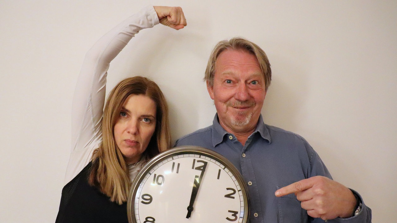 Tina Voß und Dietmar Wischmeyer halten eine Uhr, die auf kurz nach 12 steht.